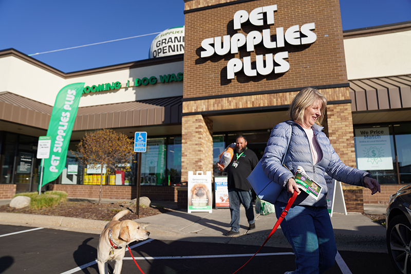 a woman leaving a pet supplies plus store with her dog on a leash. An employee follows carrying her purchases