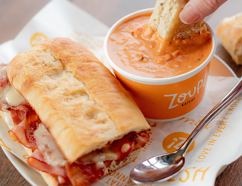 A hot sandwich next to a cup of tomato soup with a piece of bread being dipped